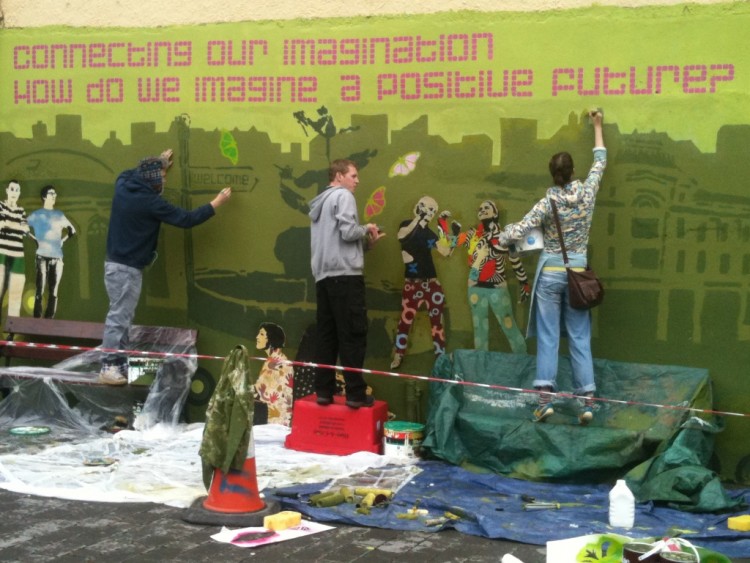 Young people painting a mural. It reads 'Connecting our imagination, how do we imagine a positive future?'